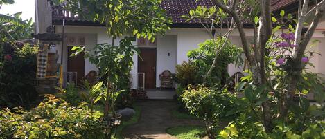 A traditional house with a tiled roof, surrounded by lush greenery and a paved pathway leading to the entrance.