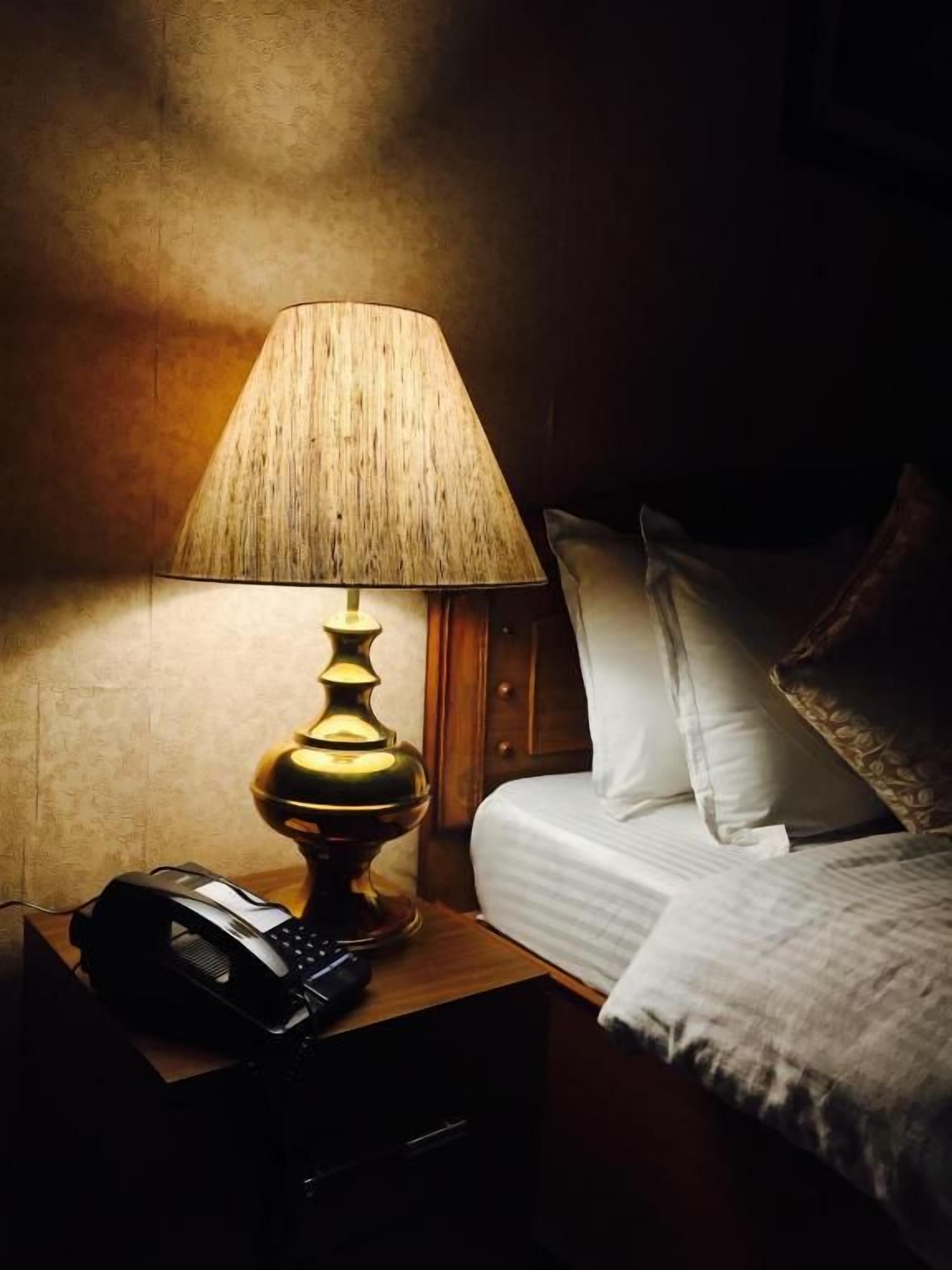 A bedside table with a lamp, a telephone, and a bed with pillows.