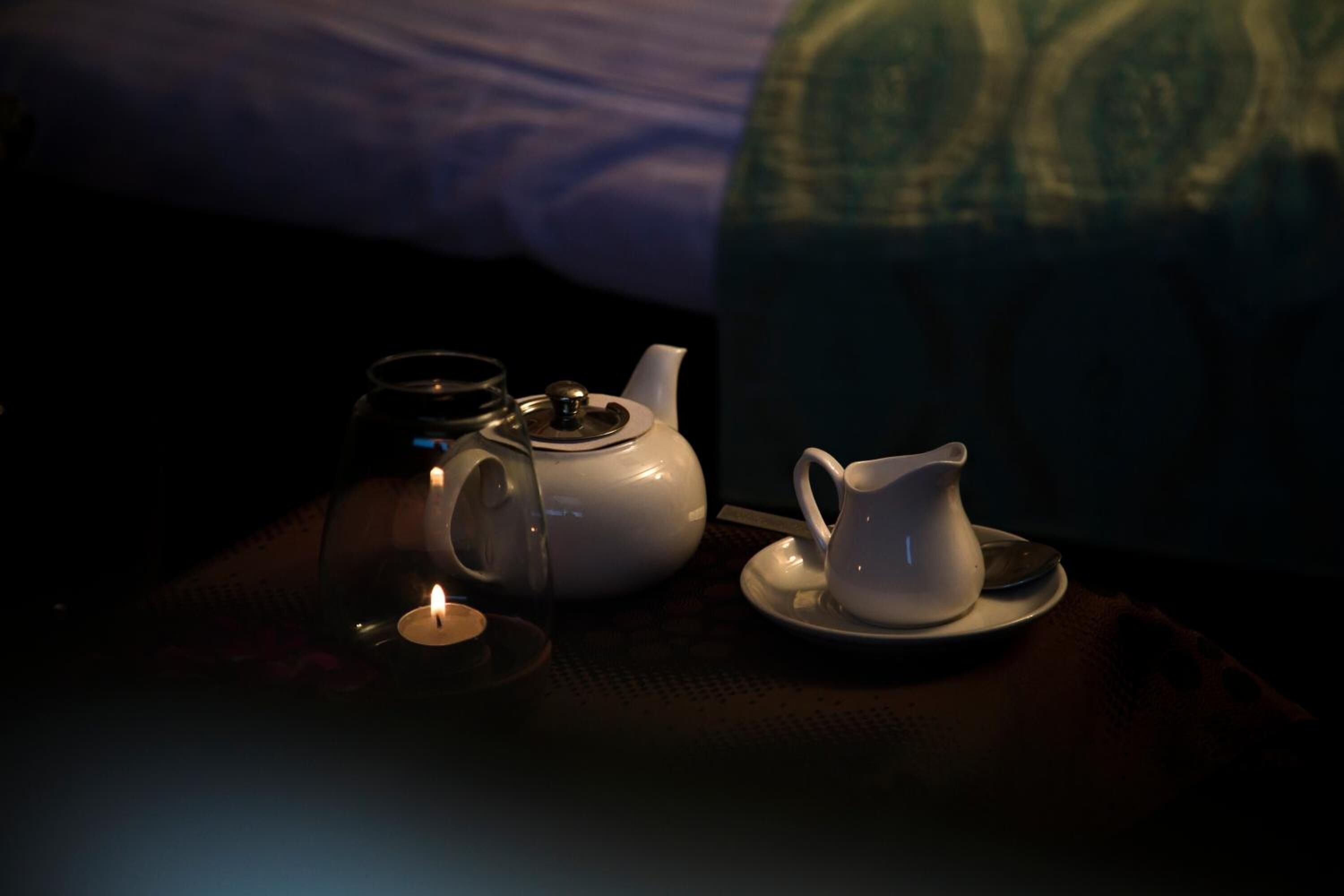 Una habitación con poca luz, una cama, una tetera blanca, una taza y una vela sobre una mesa.