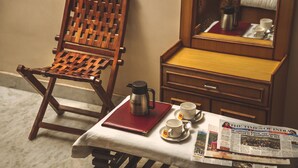A wooden chair, a table with a red notebook, a coffee maker, cups, and a newspaper. - Hotel Meenakshi Udaipur (Udaipur)