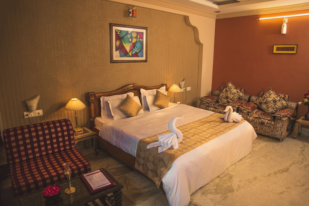 Une chambre d’hôtel avec un grand lit, un canapé à carreaux, une petite table avec un vase et une lampe fixée au mur.