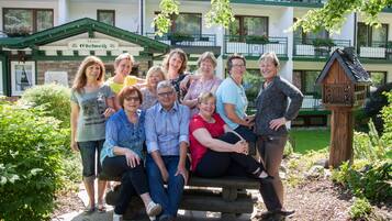Een groep mensen poseert voor een hotel met een groen en wit gevel, een houten bankje en een vogelhuisje in de tuin.