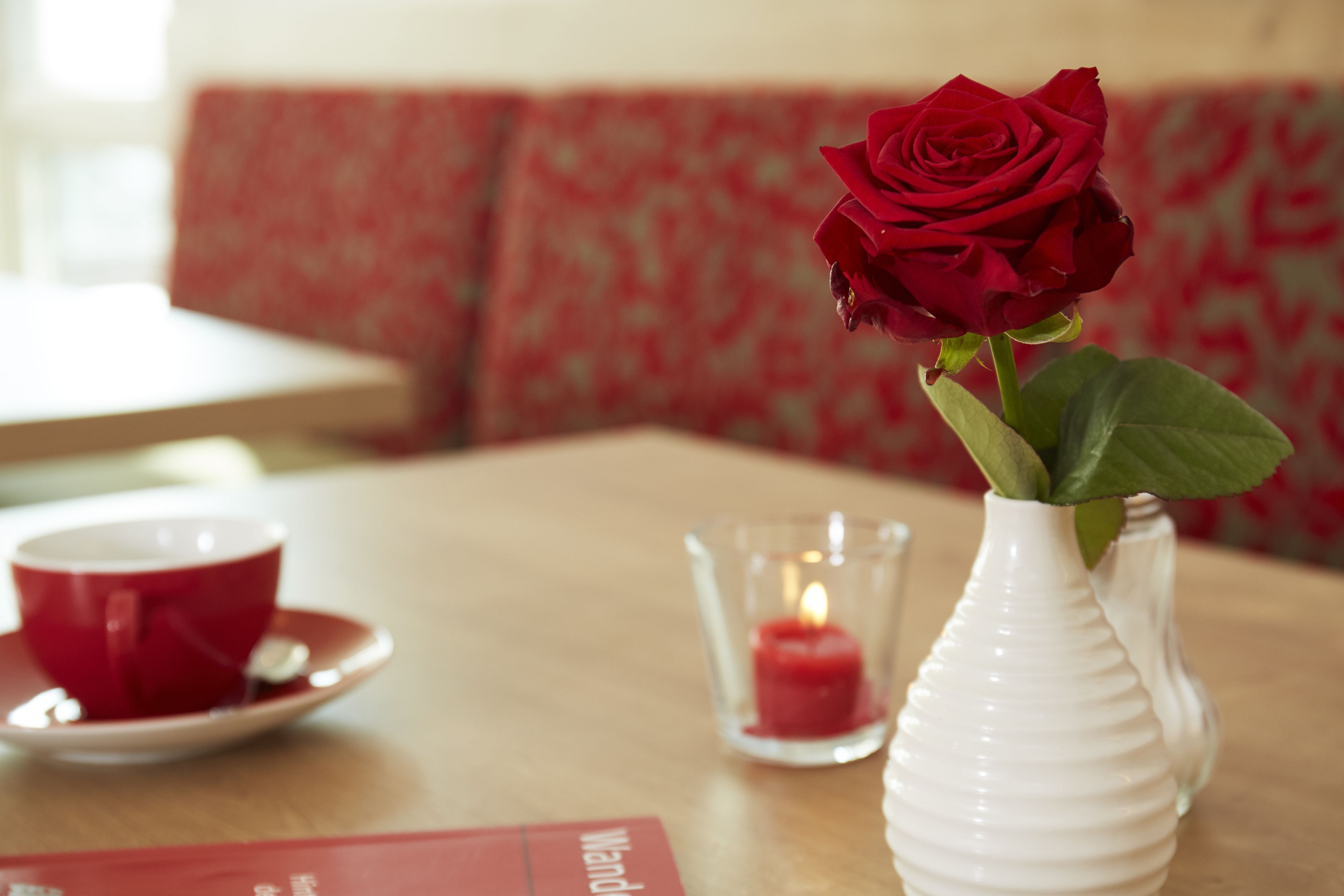 Una mesa con un jarrón blanco que contiene una rosa roja, una taza y platillo rojos, y una vela encendida en un candelabro de vidrio.