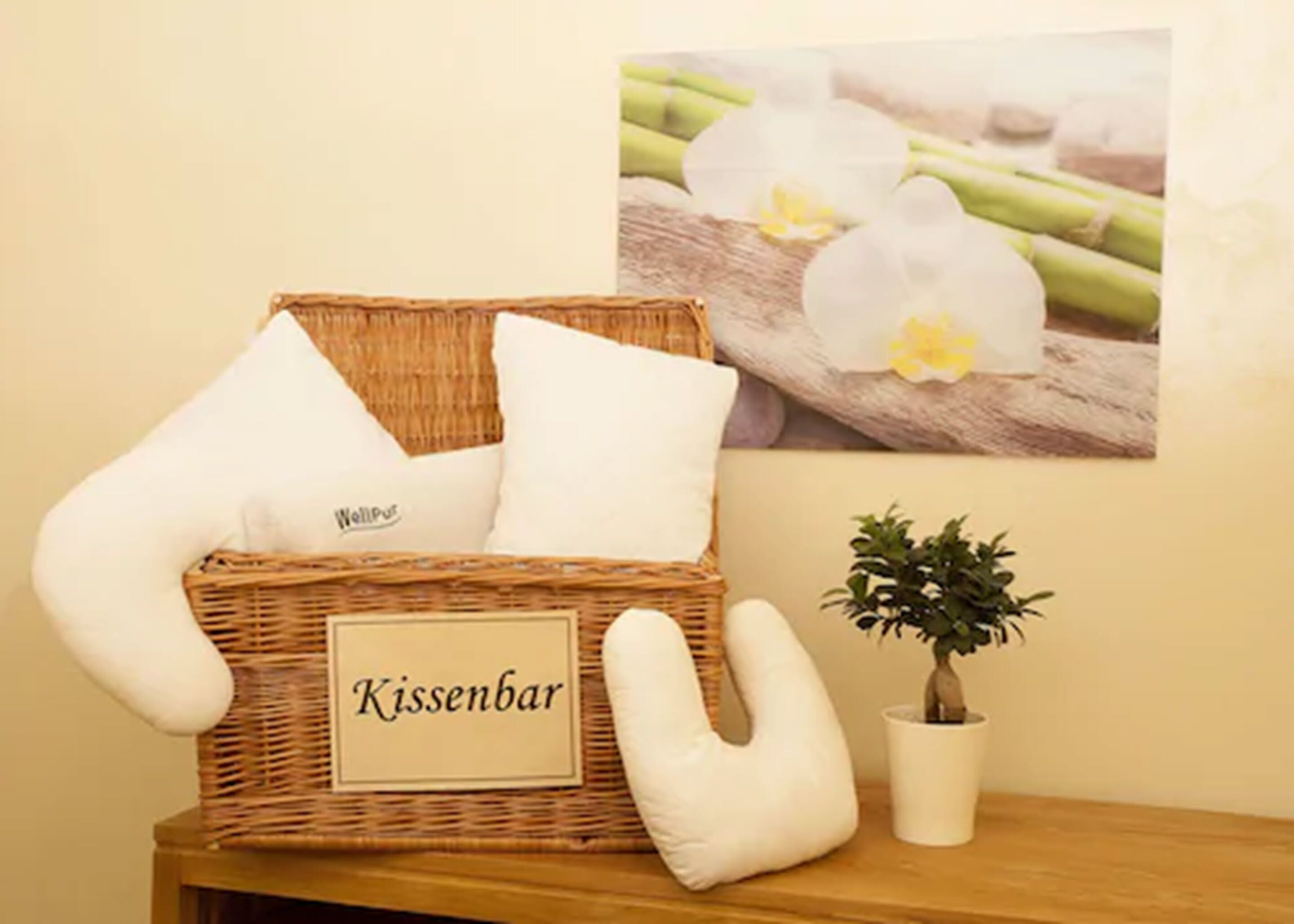 A wicker basket labeled "Kissenbar" with white cushions, a small potted plant, and a framed picture of flowers on a wooden surface.