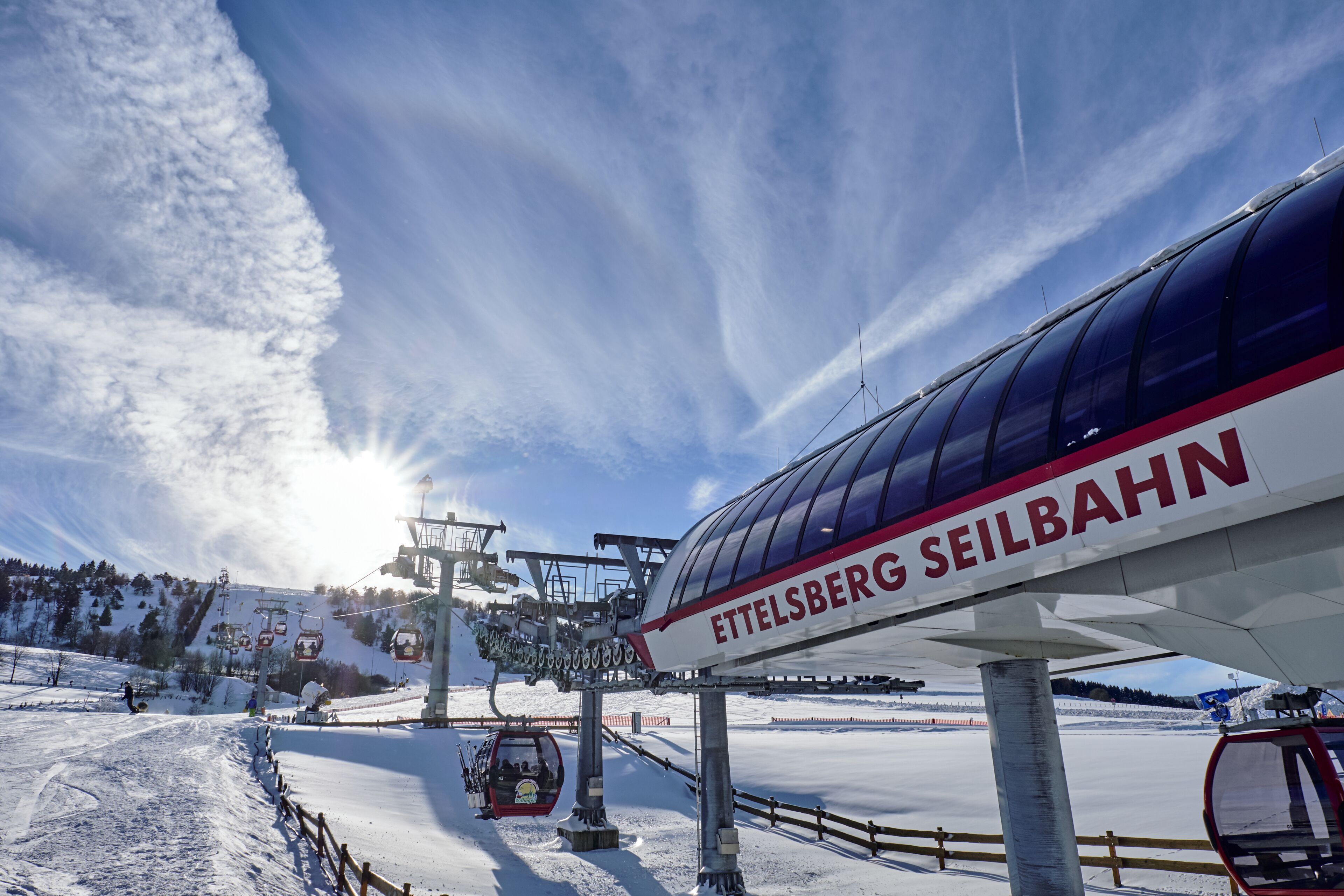 A ski lift with the name "ETTELSBERG SEILBAHN" and a snowy landscape with a clear sky.