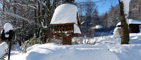 A wooden birdhouse covered in snow, surrounded by snow-covered trees and a clear blue sky.
