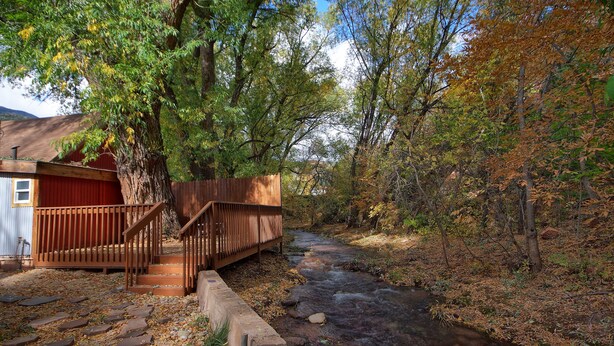 A rustic cabin with a wooden staircase leading to a red door, surrounded by a forested area and a flowing stream.