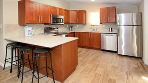 A modern kitchen with wooden cabinets, a stainless steel refrigerator, a microwave, and a coffee maker. There are three black bar stools at the island.