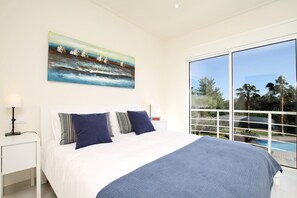 A bedroom with a large bed, a painting on the wall, and a view of a pool through the window.