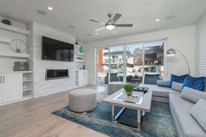 A modern living room with a flat-screen TV, a ceiling fan, a sofa, ottomans, and a coffee table. - Beachside Retreat with Rooftop Deck (Newport Beach)