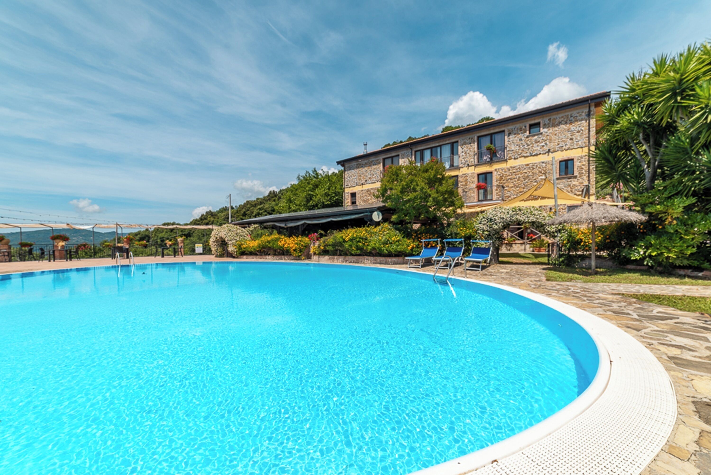 A swimming pool with clear blue water, surrounded by a stone deck. There are lounge chairs and a building with a stone facade in the background.