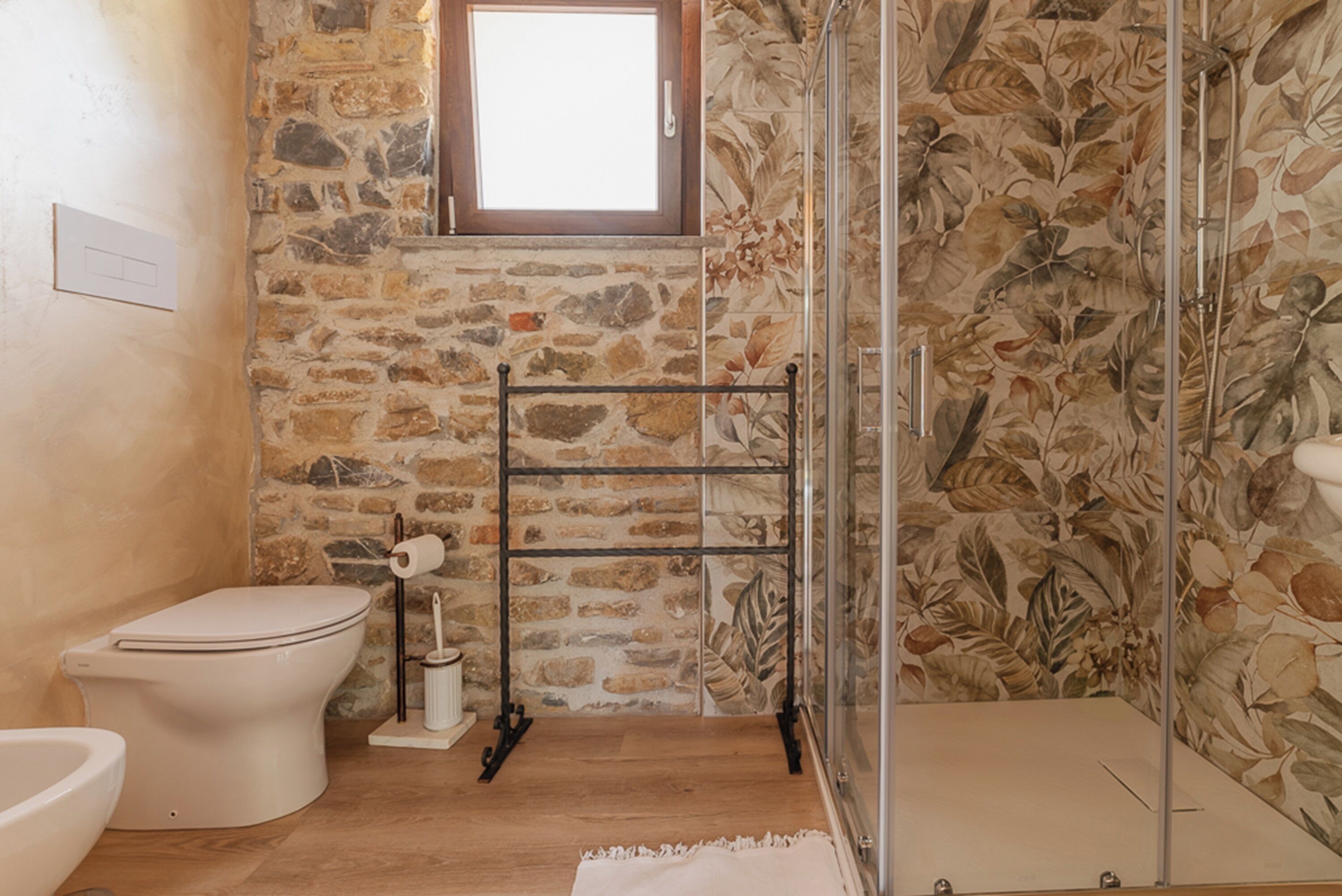 A bathroom with a stone wall, a toilet, a shower with a glass door, and a wooden floor.