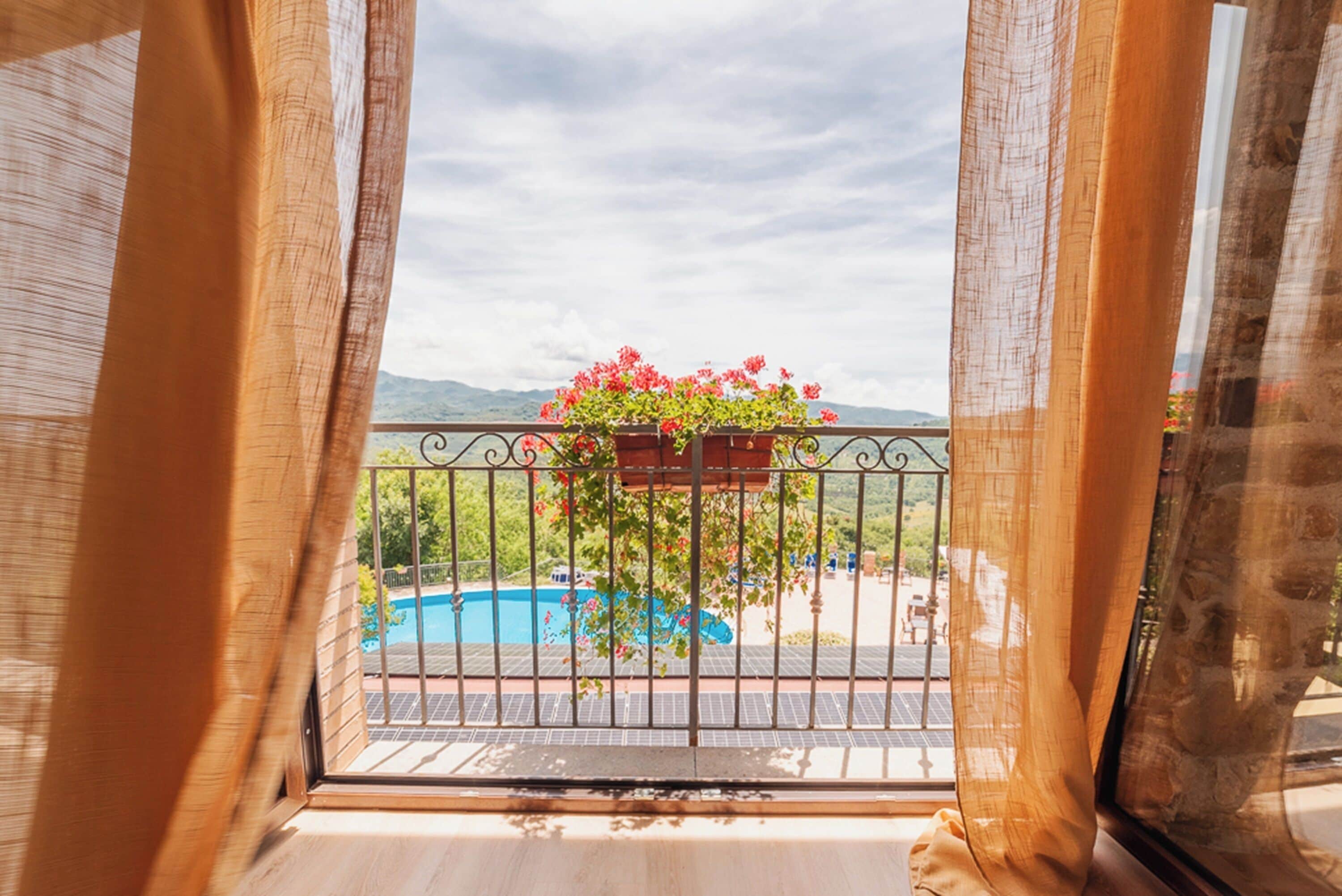 Un balcón con barandilla de metal, una maceta colgante con flores rojas, y una vista de una piscina y montañas al fondo.