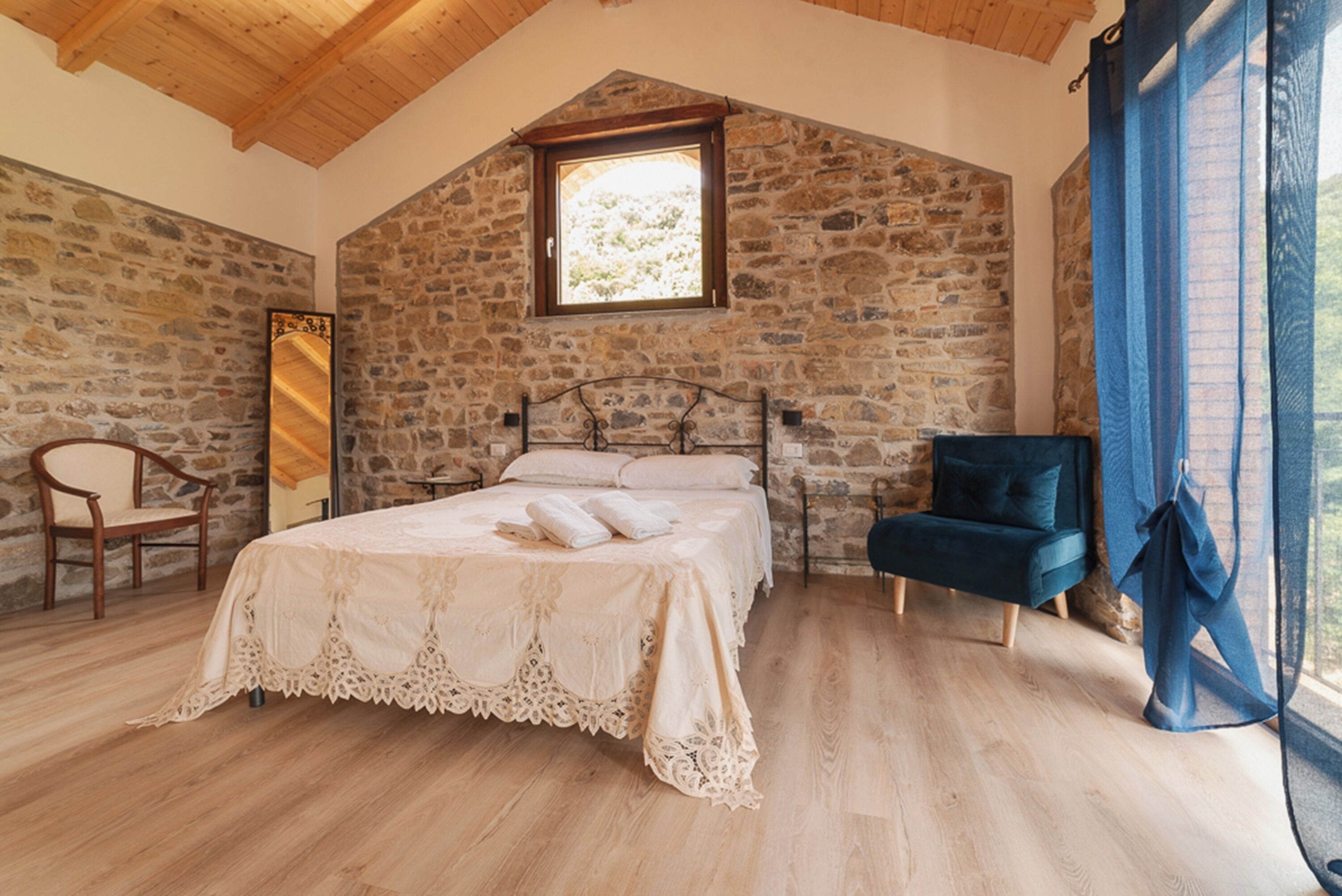 Un dormitorio con pared de piedra, techo de madera, cama con cubrecama blanco, una silla y ventana con vistas al exterior.
