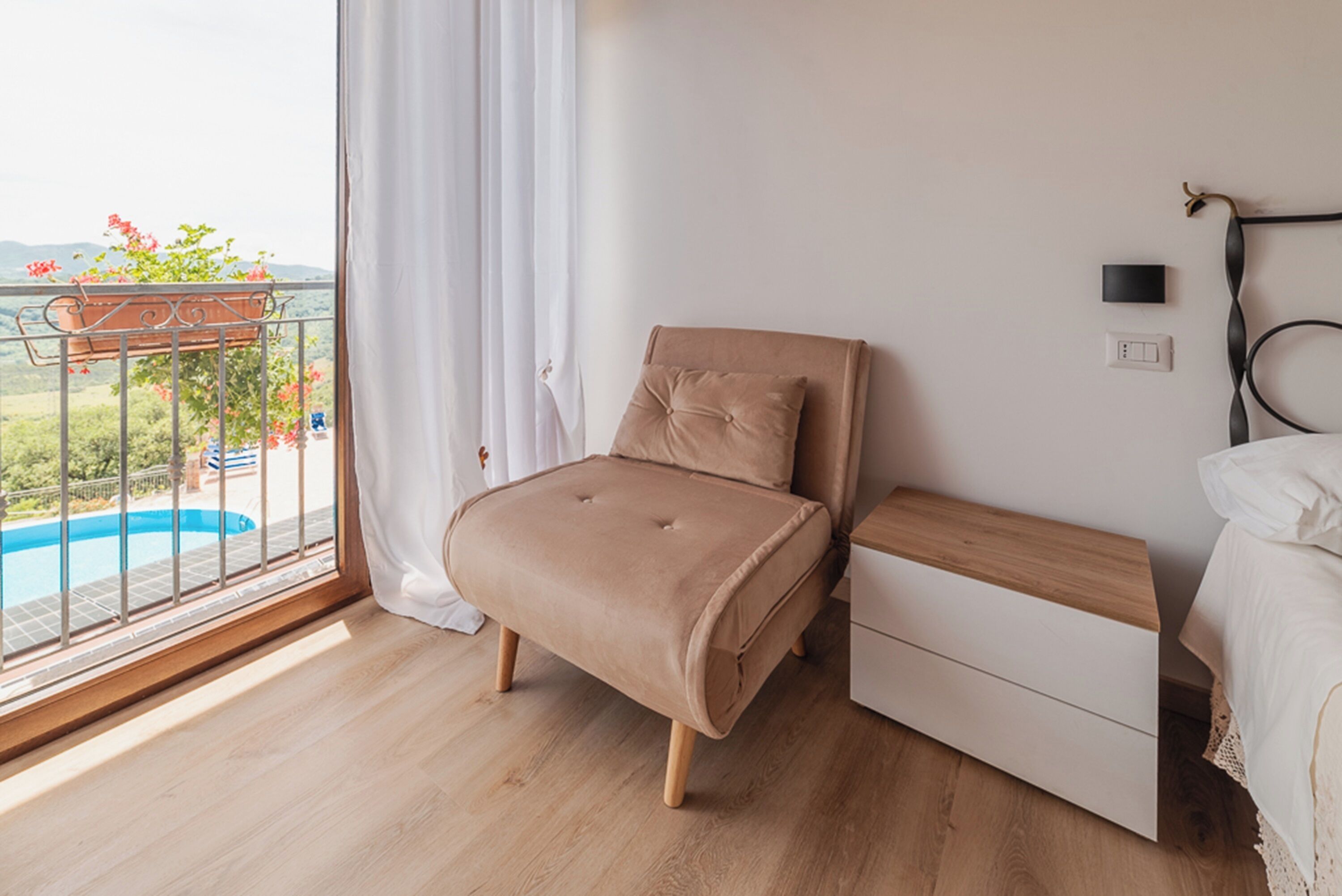 Ein Schlafzimmer mit einem Bett, einem Nachttisch, einer Holzkommode und Blick auf einen Swimmingpool vom Balkon aus.