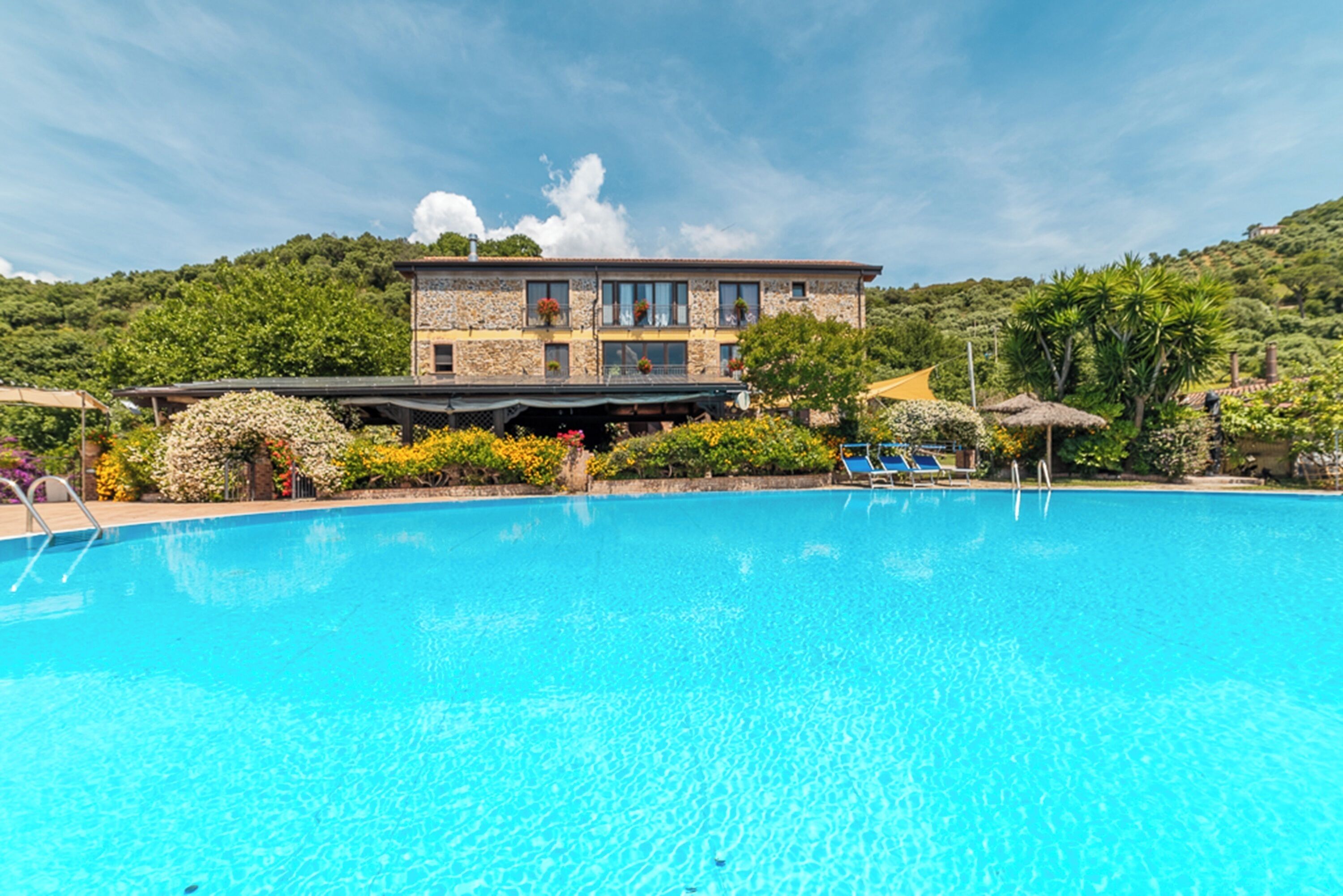 Ein Schwimmbecken mit einem dahinter liegenden Steinhaus, umgeben von viel Grün und blauem Himmel.