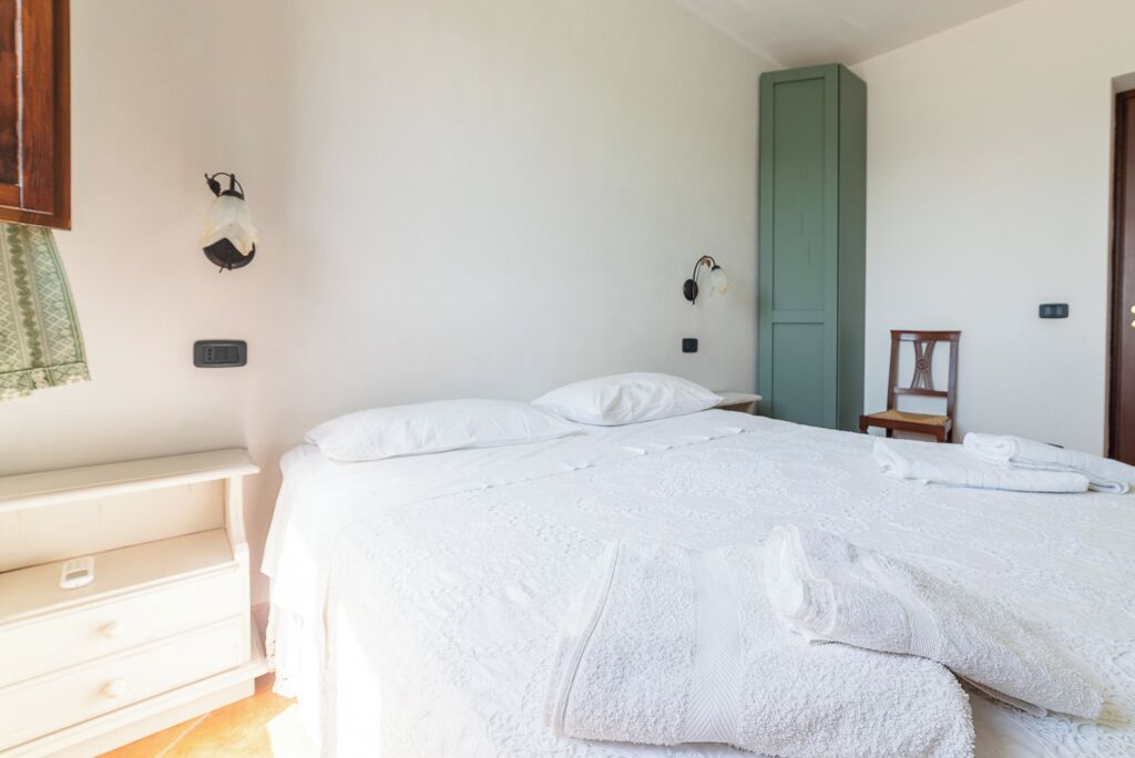 Una cama bien hecha con sábanas y toallas blancas, una mesita de noche, una lámpara de pared y una puerta verde.