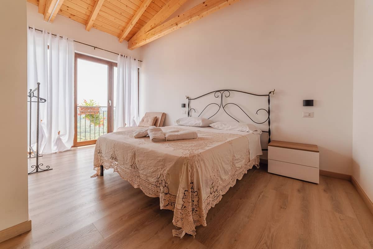 Un dormitorio con techo de madera, una cama con cubrecama de encaje, una mesita de noche y un ventanal corredero que da a un balcón.