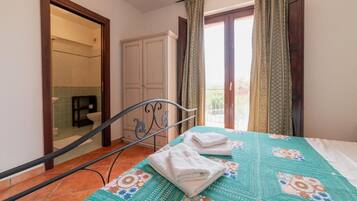 Un dormitorio con cama, armario, baño visible a través de una puerta abierta y balcón con cortinas.