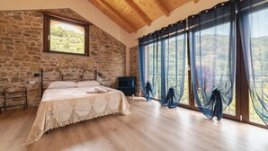 Un dormitorio con pared de piedra, techo de madera, una cama, un televisor y ventanas grandes con cortinas.