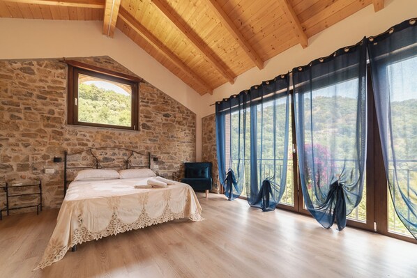 Un dormitorio con pared de piedra, techo de madera, una cama, un televisor y ventanas grandes con cortinas.