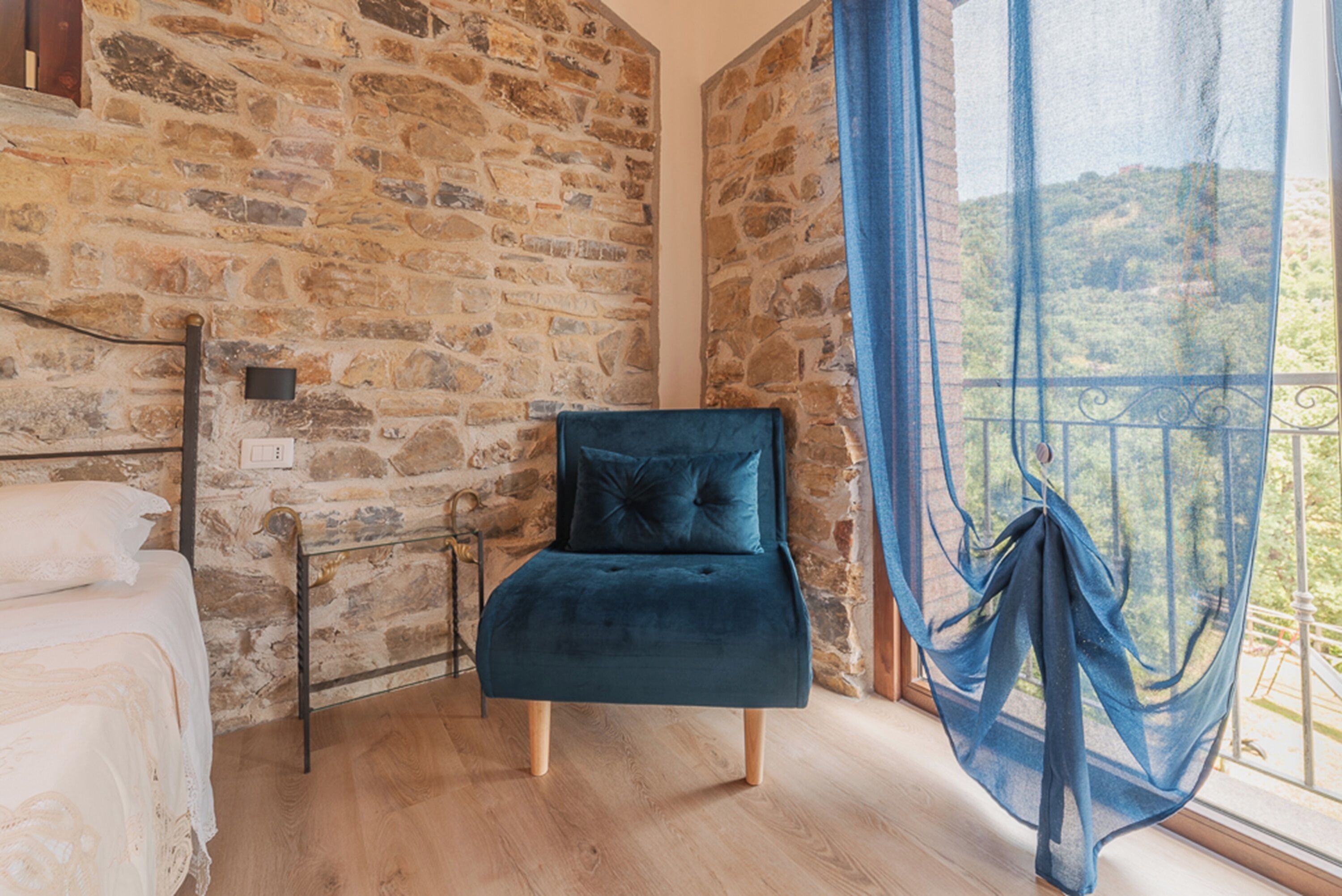 Ein Zimmer mit Stehwänden, einem Bett, einem blauen Sessel und einem Fenster mit Blick auf eine Hügellandschaft.