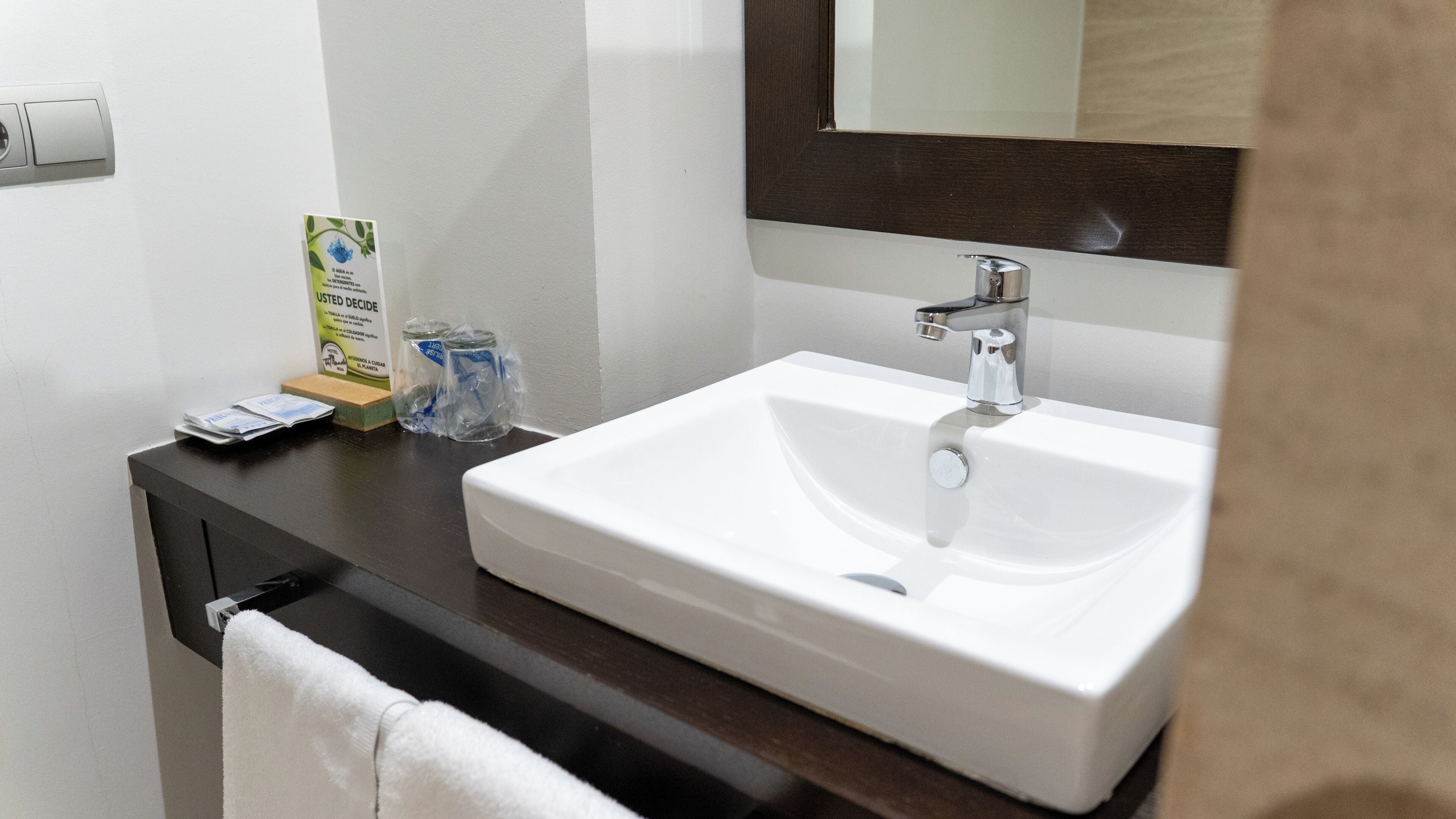 Une salle de bain avec un lavabo blanc, un miroir, une serviette et une petite boîte posée sur le comptoir.