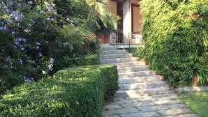 Una escalera de piedra que conduce a la entrada de un edificio, rodeada de setos cuidados y plantas con flores.