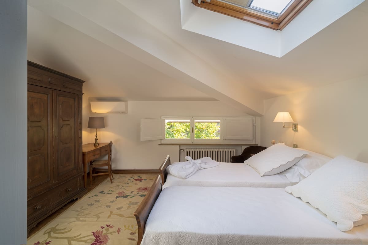 Ein Schlafzimmer mit zwei Betten, einem Holzschrank, einem Schreibtisch mit Lampe, einem Heizkörper und einem Oberlicht.