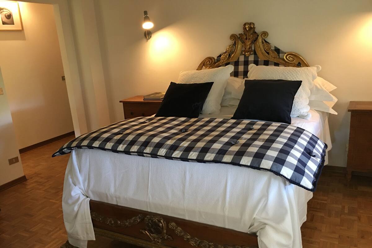 Ein Bett mit kariertem Überwurf, ein Holzkopfende mit floralen Schnitzereien und ein Nachttisch mit einer Lampe.
