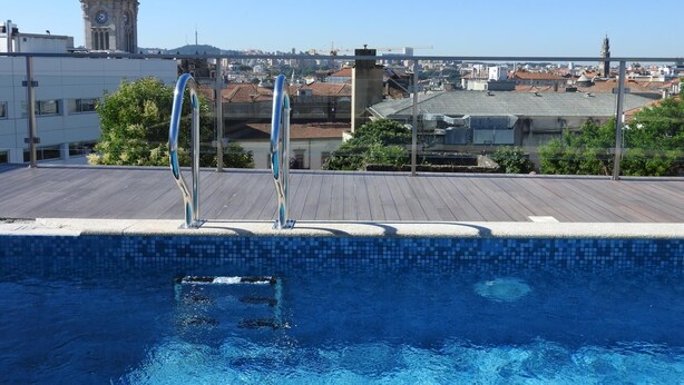 A rooftop pool with clear blue water, surrounded by a metal railing. In the background, there is a clock tower and a cityscape with various buildings.