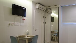 A hotel room with a bed, a TV mounted on the wall, a small table with chairs, an air conditioning unit, and a sliding glass door leading to a balcony.