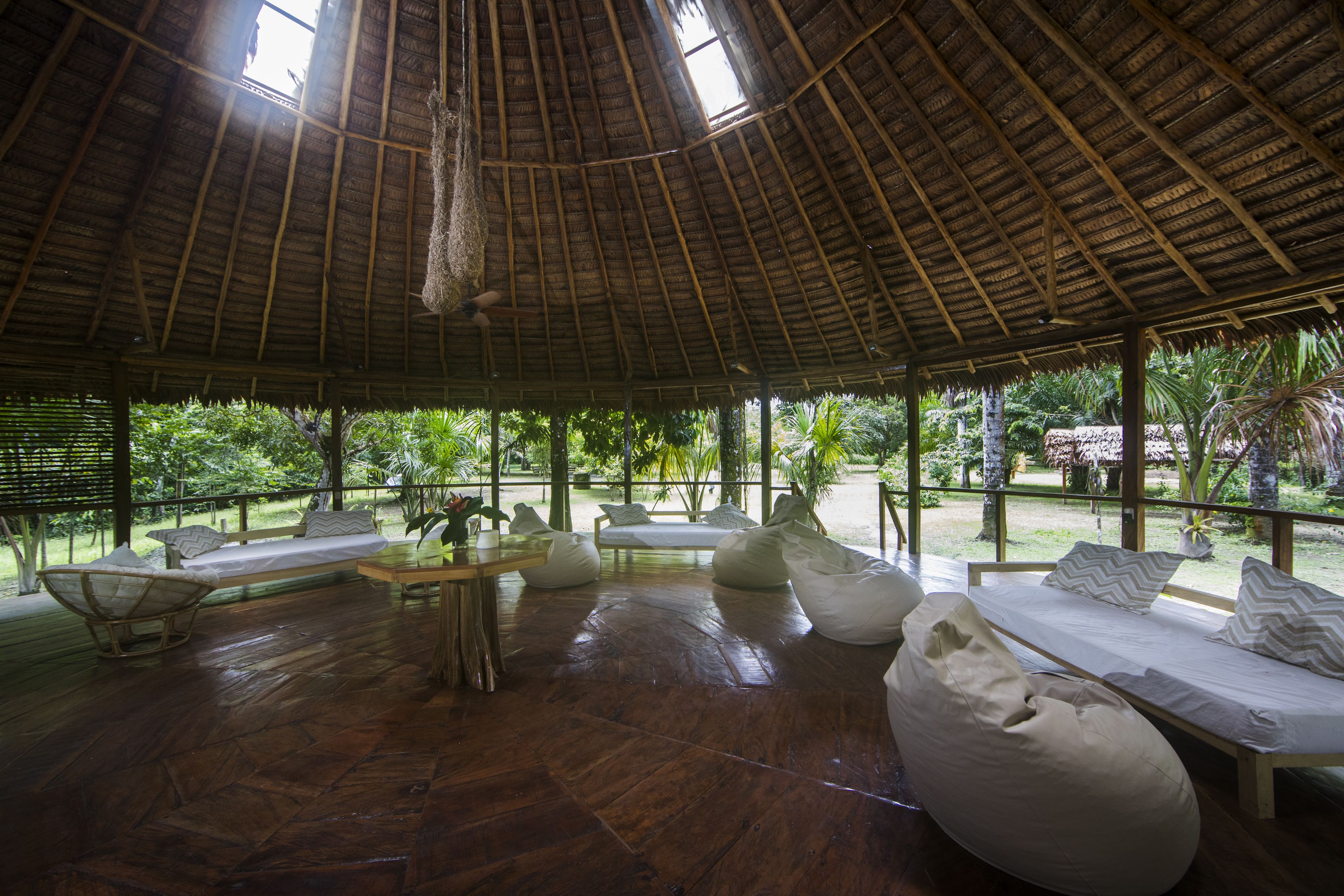 Un salón al aire libre con suelo de madera, techo de paja y vista a exuberante vegetación.