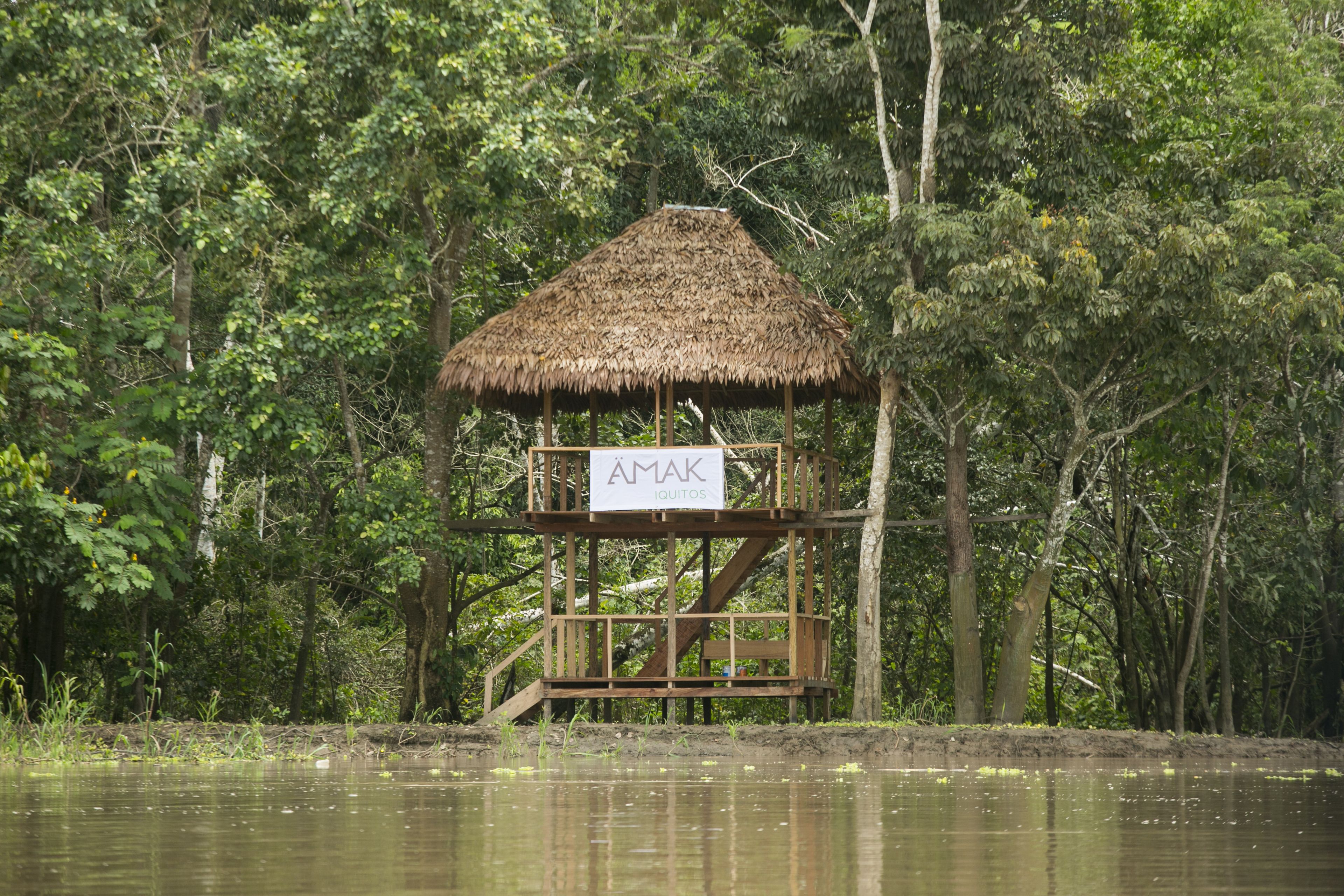 Une structure en bois, surmontée d’un toit de chaume, portant l’inscription « AMAK IQUITOS », située dans une zone boisée près de l’eau.