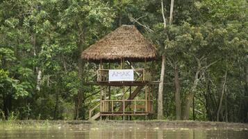 Une structure en bois, surmontée d’un toit de chaume, portant l’inscription « AMAK IQUITOS », située dans une zone boisée près de l’eau.