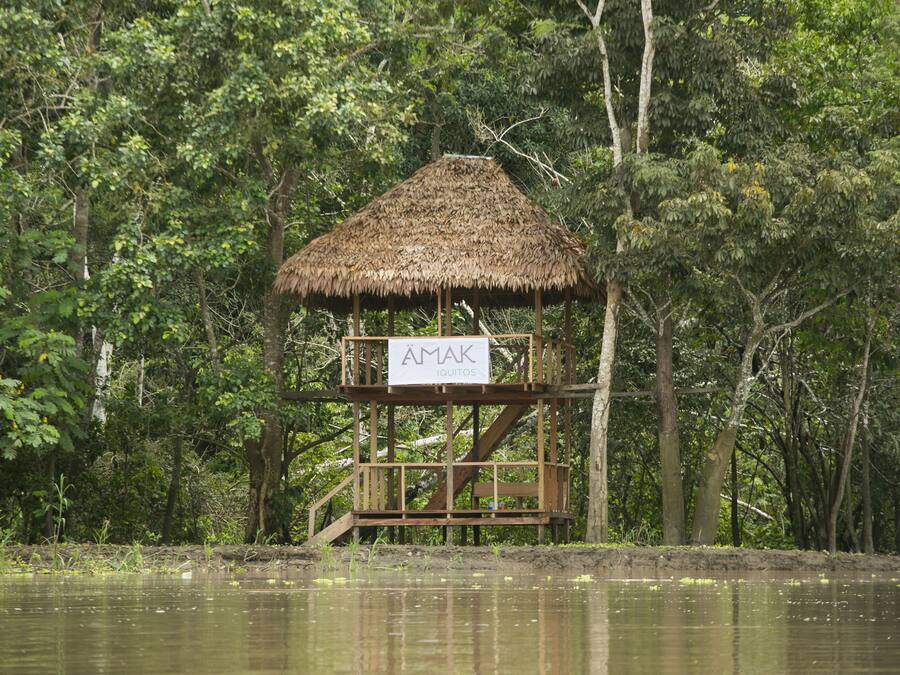 Une structure en bois, surmontée d’un toit de chaume, portant l’inscription « AMAK IQUITOS », située dans une zone boisée près de l’eau.