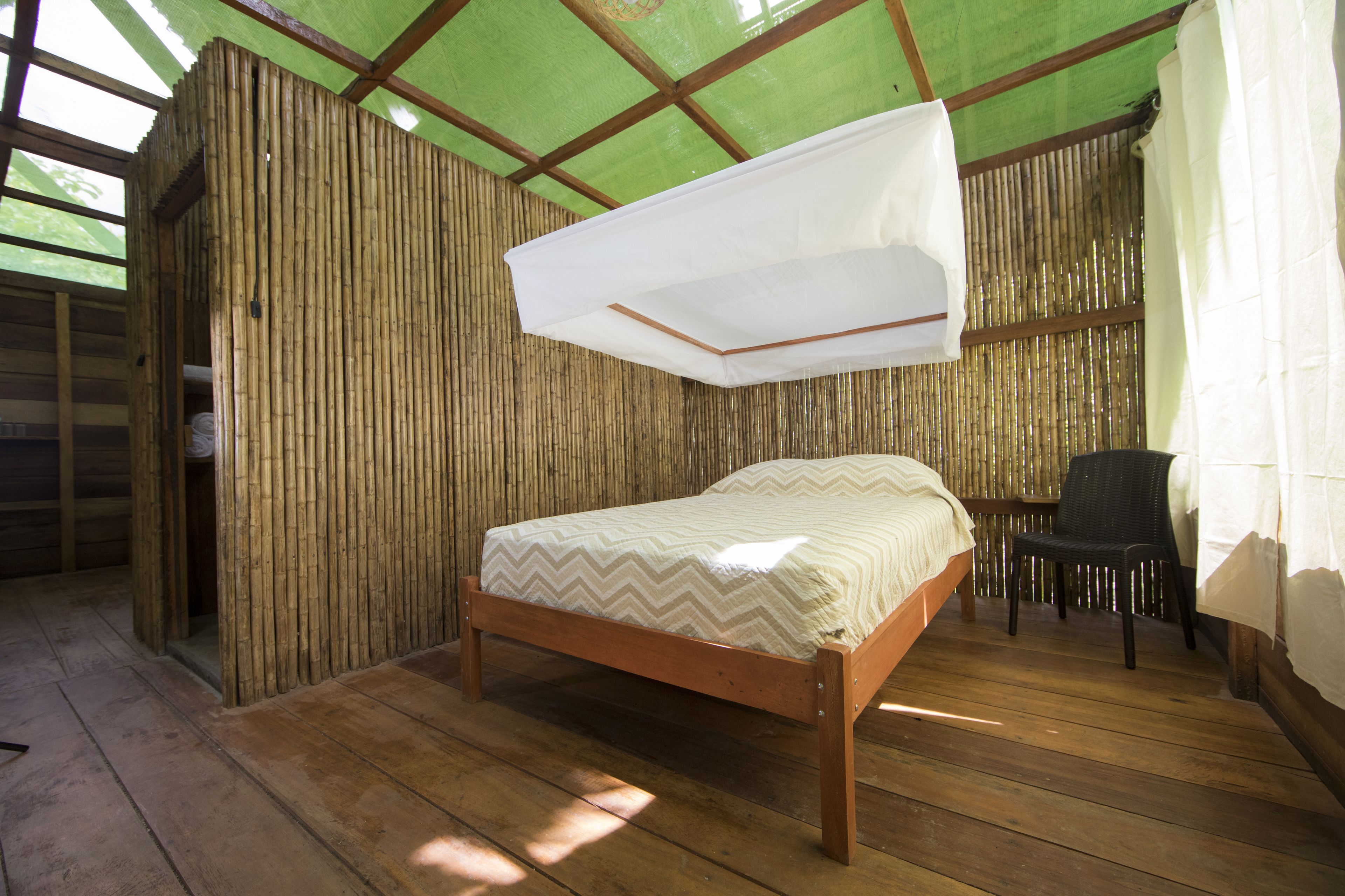 Una cama de madera con mosquitero blanco, una silla y paredes de bambú en una habitación con suelo de madera.