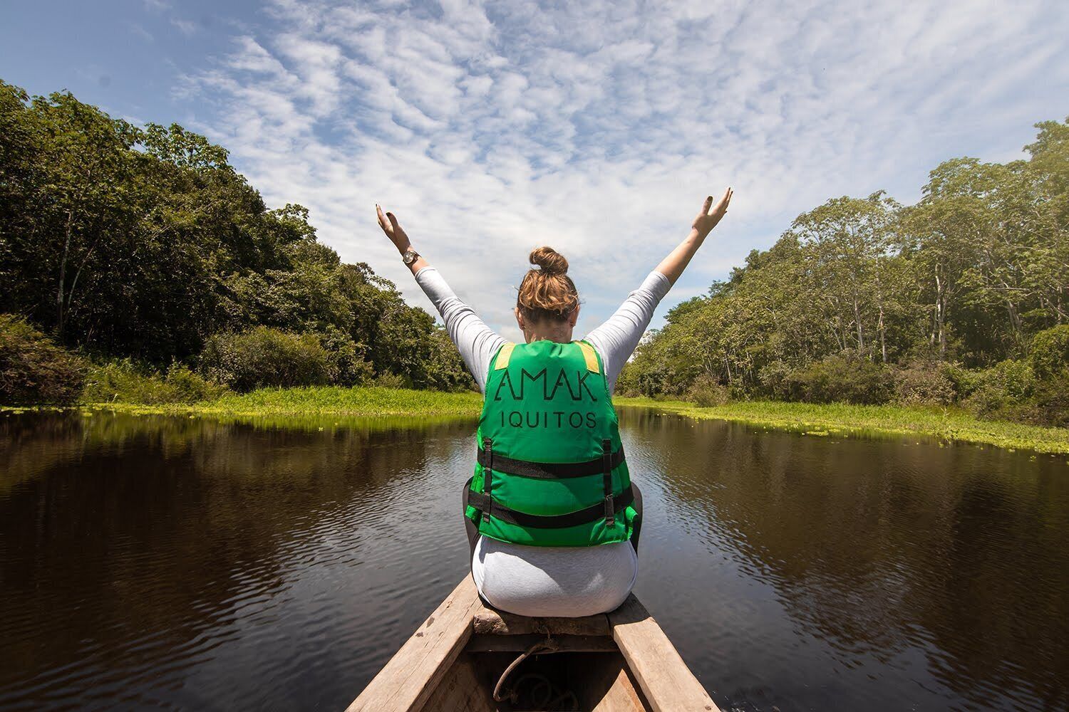 Una persona en una canoa, usando chaleco salvavidas verde con la inscripción "AMAK IQUITOS" en la parte posterior, rema por un río tranquilo rodeado de exuberante vegetación.