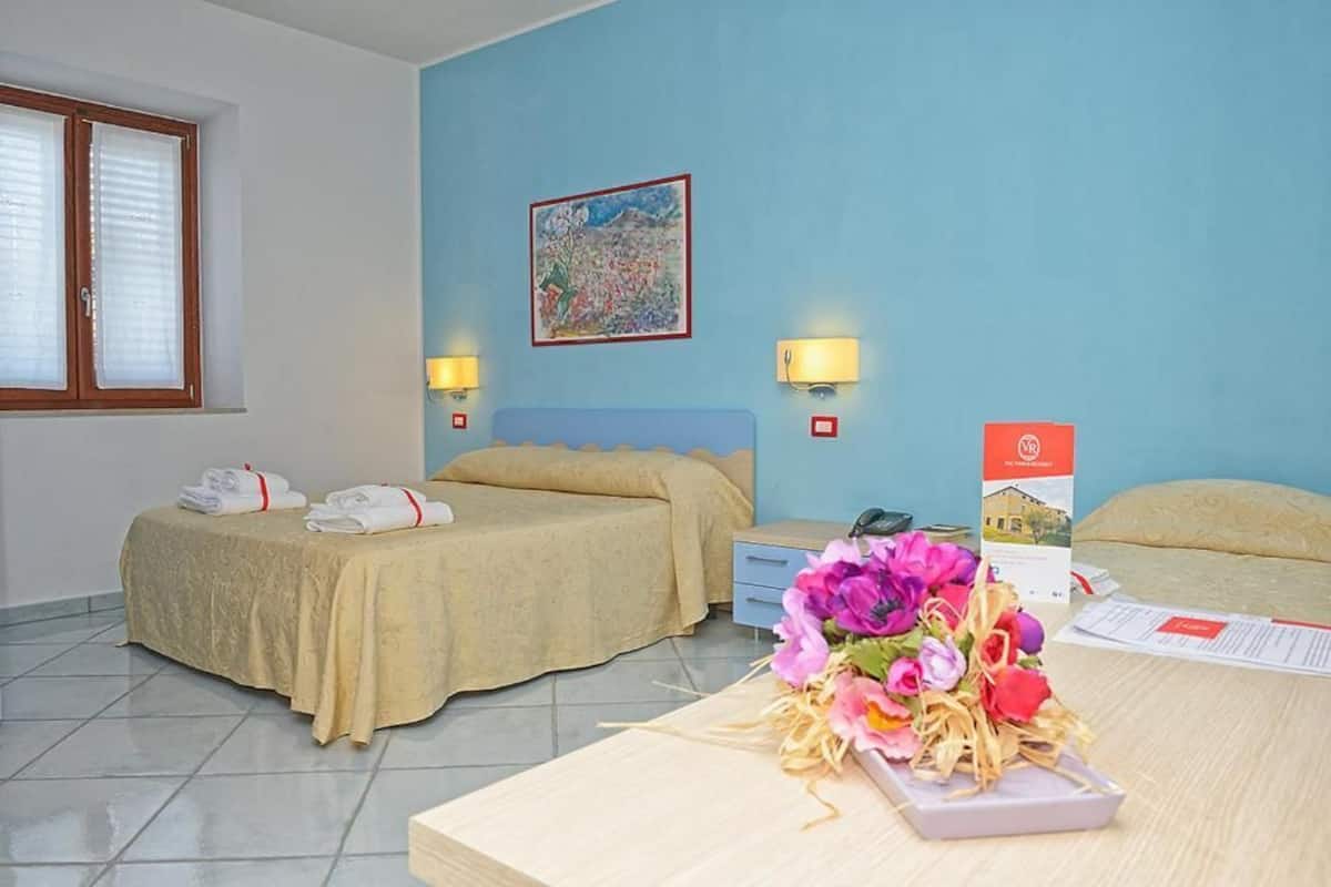 A hotel room with two beds, a desk, a nightstand, a window with blinds, a painting on the wall, and a bouquet of flowers on the desk.