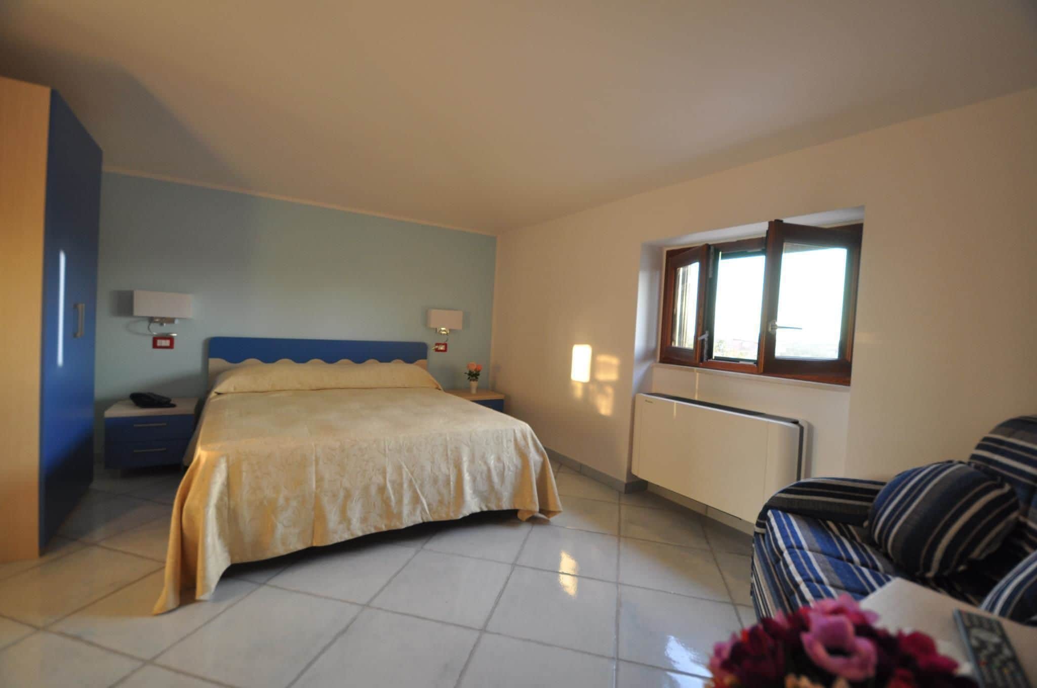A bedroom with a bed, a nightstand, a lamp, a window, and a sofa.