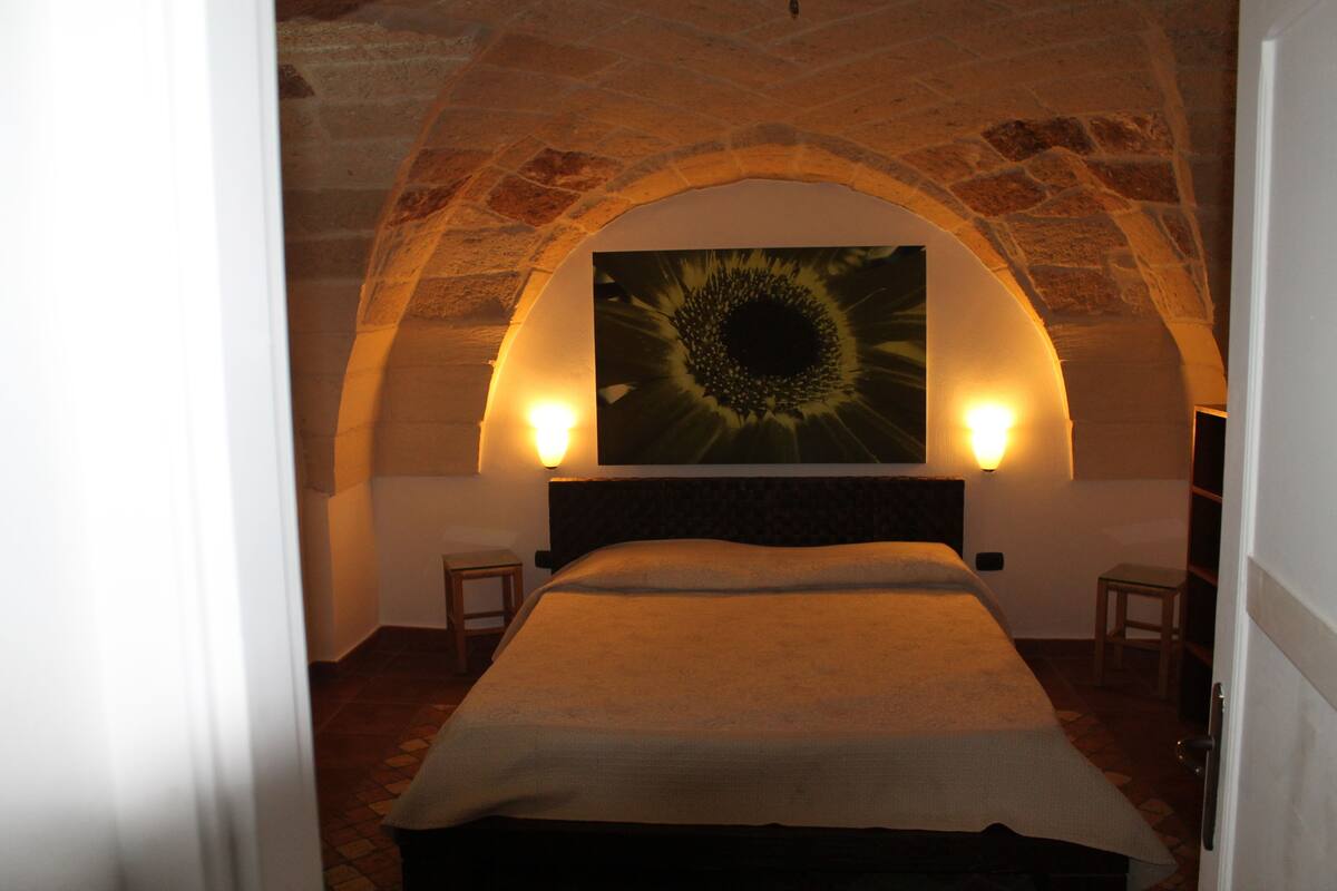 Quarto com teto abobadado em pedra, uma cama grande com cabeceira, duas mesas de cabeceira e um candeeiro fixado na parede.