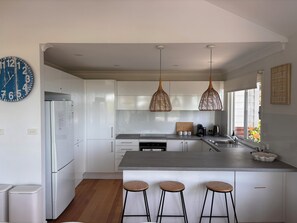 A modern kitchen with white cabinetry, a central island with stools, pendant lights, and a large wall clock. - Beach House for Family or Couples with views & 2 minute walk to beach and shops (Copacabana)