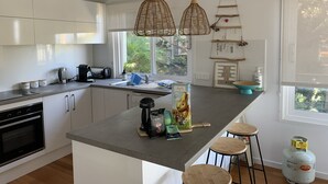 Eine moderne Küche mit weißen Schränken, einem Einbauofen, einer Kaffeemaschine und einem Gasherd. Zwei hängende Weidenkorb-Pendelleuchten, ein Fenster mit Blick auf Grünflächen und eine Gasflasche auf dem Boden.