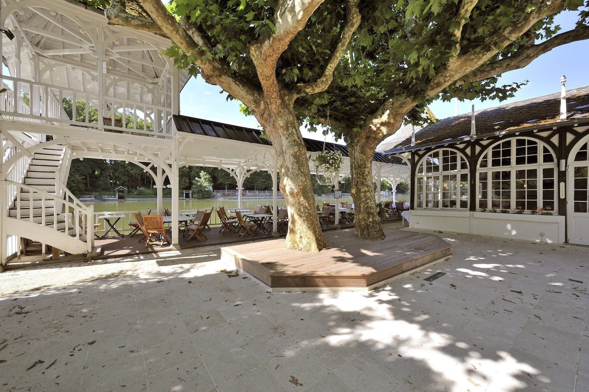 A covered outdoor seating area with tables and chairs, a staircase, and a large tree providing shade.