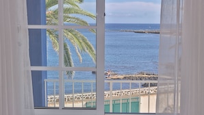 Une vue à travers une fenêtre à cadre blanc, donnant sur une étendue d’eau, une côte rocheuse et un palmier.