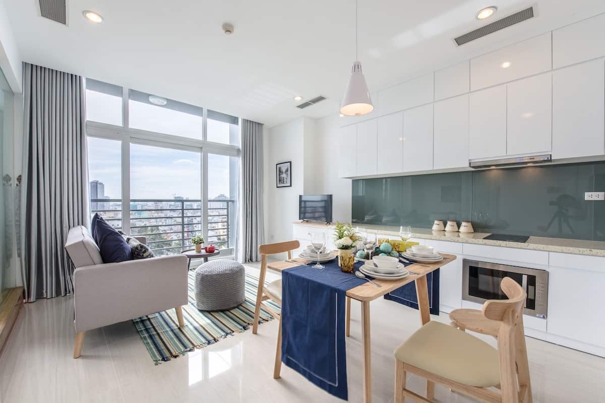 A modern apartment with a dining area, kitchen, and a large window offering a city view.