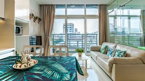 A modern living room with a sofa, a dining table with a tropical tablecloth, a small potted plant, and a view of the city through large windows. - Luxury Ben Thanh TAA Apartments (Ho Chi Minh City)