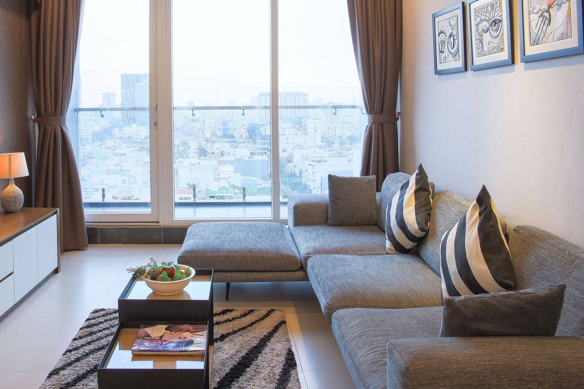 A modern living room with a large flat-screen TV, a sofa, a coffee table with a bowl of fruit, and a view of a cityscape through large windows.