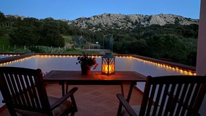 Zona junto a la piscina con dos sillas de madera, una mesa con planta en maceta y un farol.