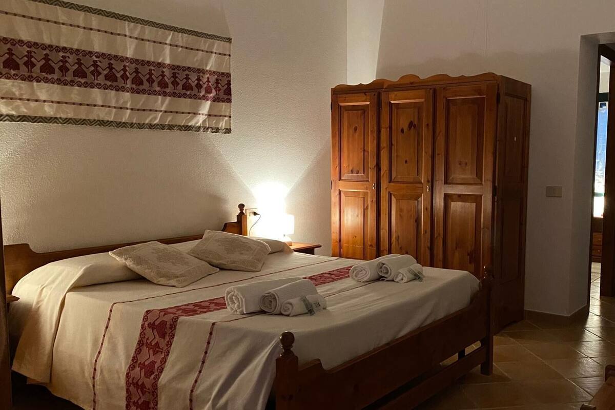 A bedroom with a wooden bed, a nightstand, a wardrobe, and a patterned tapestry on the wall.